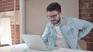 Homem jovem trabalhando de frente ao seu laptop com dores nas costas: isso pode ser derivado de uma má postura.